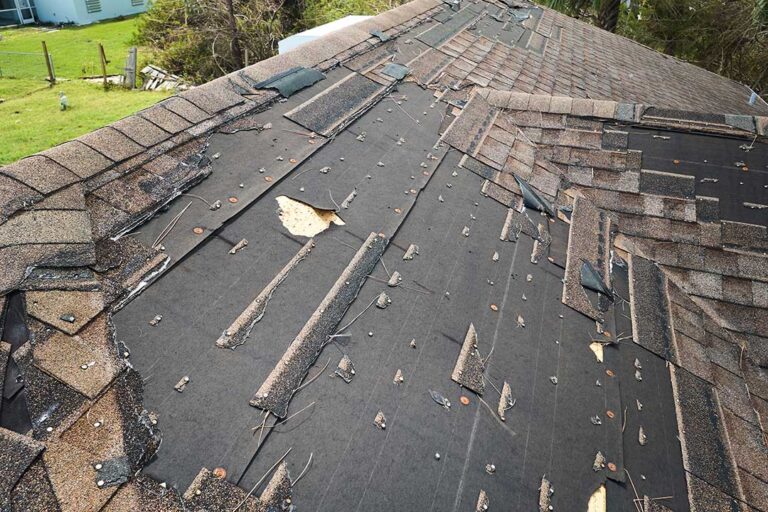 Homeowner compares roof shingle samples beside a house with light siding and snow-covered trees.